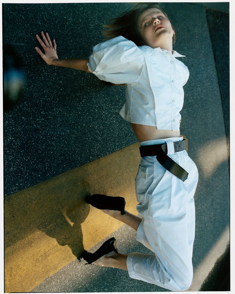 Fashion shot of a stylish woman in a white blouse and pants lying on an outdoor stone surface with her shadow visible in daylight.