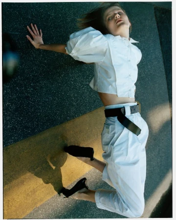 Fashion shot of a stylish woman in a white blouse and pants lying on an outdoor stone surface with her shadow visible in daylight.