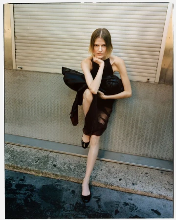 Fashion shot of a young woman in a dark dress leaning against a textured metal wall, featuring cool tones and natural outdoor lighting.