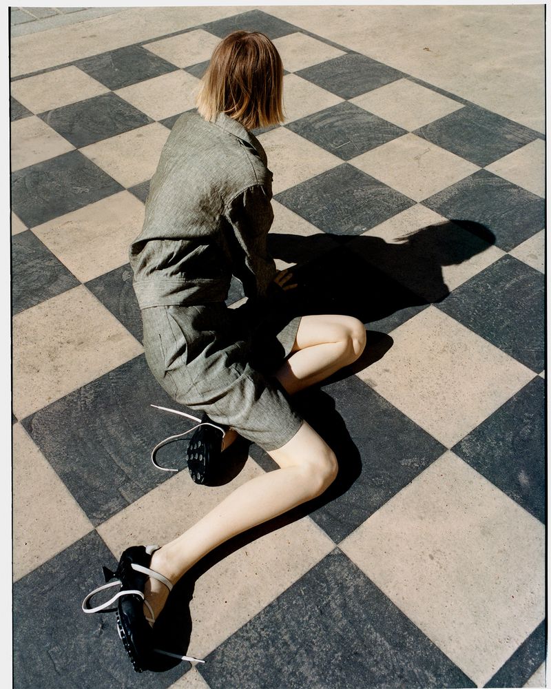 Back view of a person in a grey short suit, kneeling on a black and white checkered surface, with black shoes, casting a shadow.