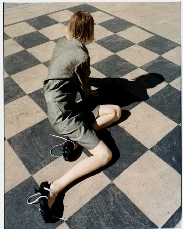 Back view of a person in a grey short suit, kneeling on a black and white checkered surface, with black shoes, casting a shadow.