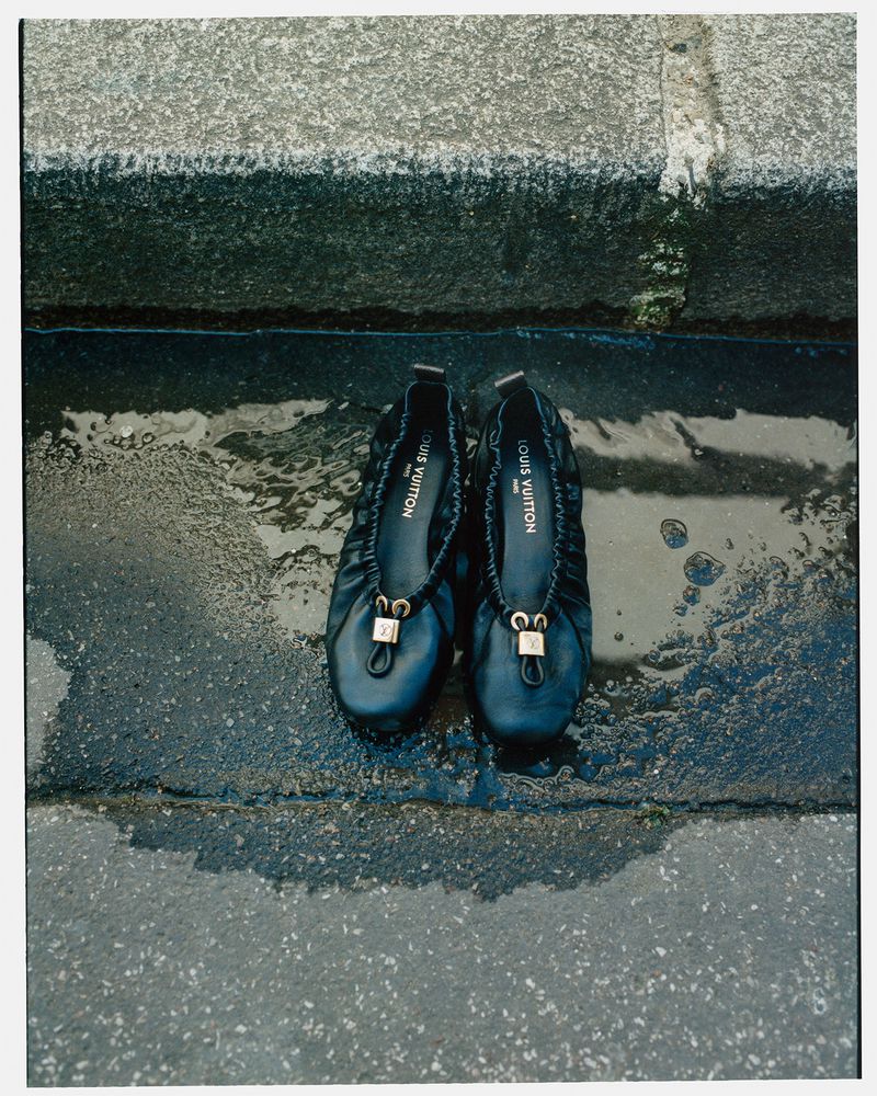 Close-up studio shot of a pair of black Louis Vuitton ballet flats sitting in a puddle of water on a stone surface, fashion-forward footwear.