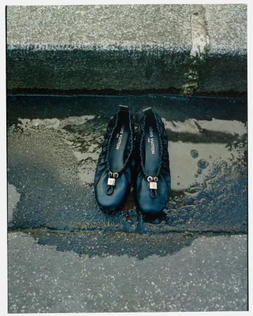 Close-up studio shot of a pair of black Louis Vuitton ballet flats sitting in a puddle of water on a stone surface, fashion-forward footwear.