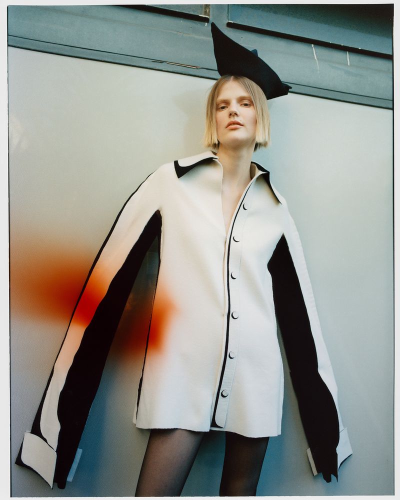 Fashion shot of a blonde model wearing an off-white jacket with black trim against a light gray background. Black hat on head.