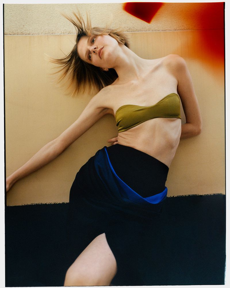 A woman poses in a bandeau top and skirt, with neutral and black background, showcasing a fashion style with minimal props and soft lighting.