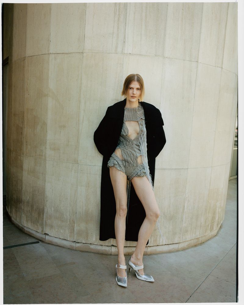 Fashion shot of a slim adult female posing in a grey, shredded bodysuit and black coat in front of a beige, curved concrete structure.