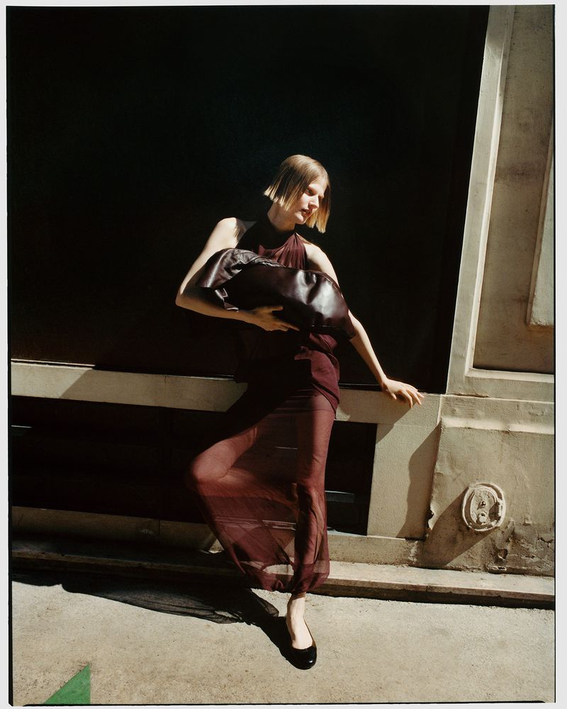 A stylish blonde woman in a sheer burgundy dress holds a large leather bag against a dark background, with natural light creating strong shadows.