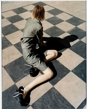 Model with short blonde hair wearing a grey co-ord set seated on a large tiled checkered ground.
