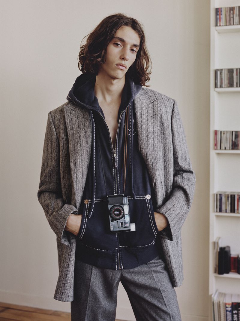 Model Nour Oitahar Normand wearing a grey patterned blazer and dark hoodie with a camera around neck in an interior setting.
