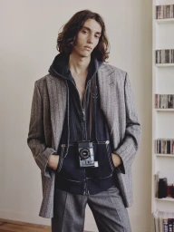 Model Nour Oitahar Normand wearing a grey patterned blazer and dark hoodie with a camera around neck in an interior setting.