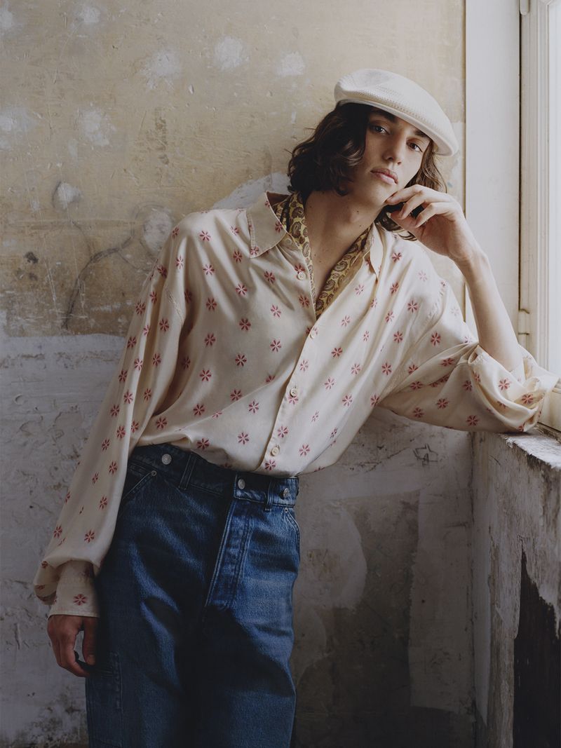 Model Nour Oitahar Normand wearing a patterned cream shirt and denim jeans leaning by a textured wall
