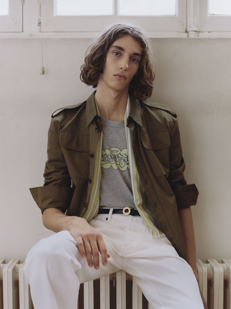 Model with long dark hair wearing an olive green jacket, graphic t-shirt, and white trousers seated against a wall