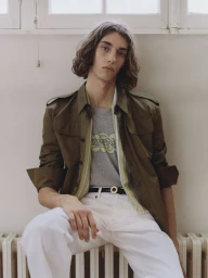 Model with long dark hair wearing an olive green jacket, graphic t-shirt, and white trousers seated against a wall