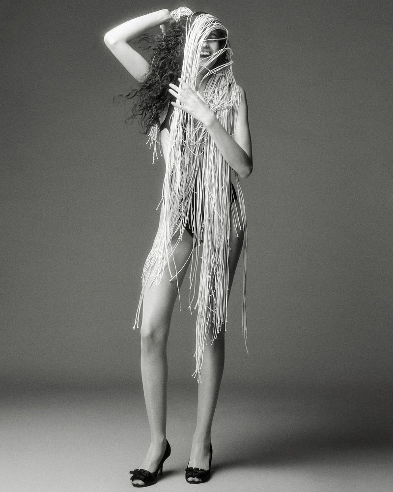 Model wearing heavily fringed garment with dark shoes against grey studio backdrop