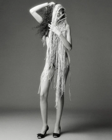 Model wearing heavily fringed garment with dark shoes against grey studio backdrop