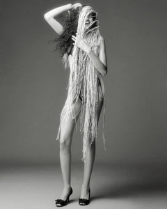 Model wearing heavily fringed garment with dark shoes against grey studio backdrop