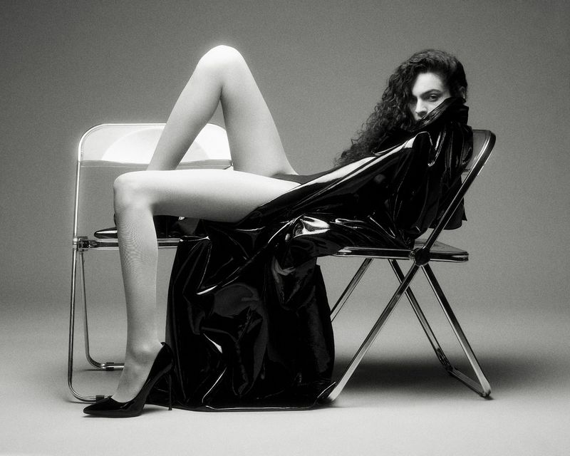 Model with long dark curly hair seated across two folding chairs wearing a glossy black trench coat and high heels