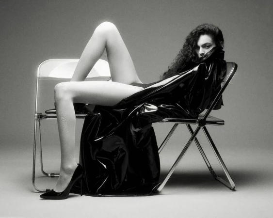 Model with long dark curly hair seated across two folding chairs wearing a glossy black trench coat and high heels