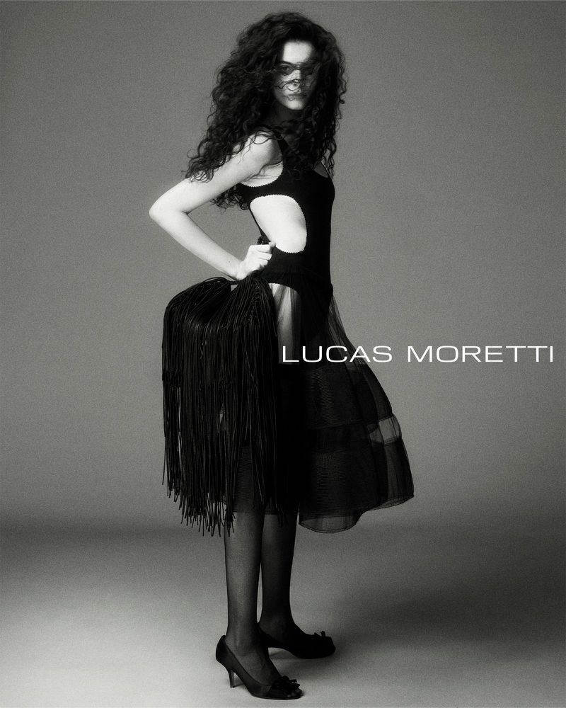 Model with long dark curly hair wearing a black cutout dress and fringed handbag against a neutral studio backdrop