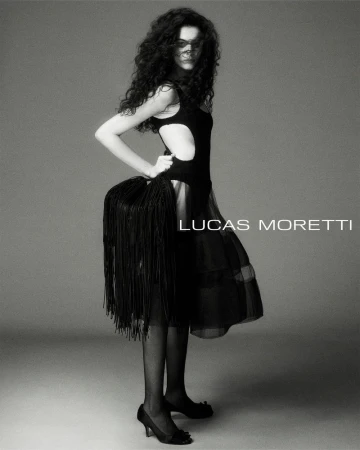 Model with long dark curly hair wearing a black cutout dress and fringed handbag against a neutral studio backdrop