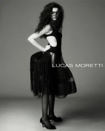 Model with long dark curly hair wearing a black cutout dress and fringed handbag against a neutral studio backdrop
