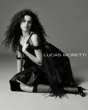 Model with long dark curly hair wearing a black dress and heels kneeling against a grey studio backdrop