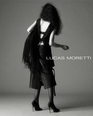 Model with long dark curly hair wearing a black sleeveless top, skirt, and high heels, carrying a fringed bag against a seamless grey backdrop.