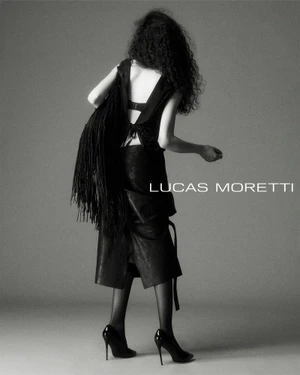 Model with long dark curly hair wearing a black sleeveless top, skirt, and high heels, carrying a fringed bag against a seamless grey backdrop.