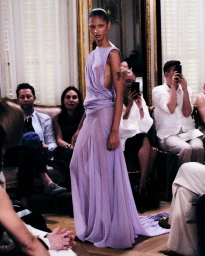 Model in a draped lilac evening gown walking down a runway before seated guests in an ornate salon.