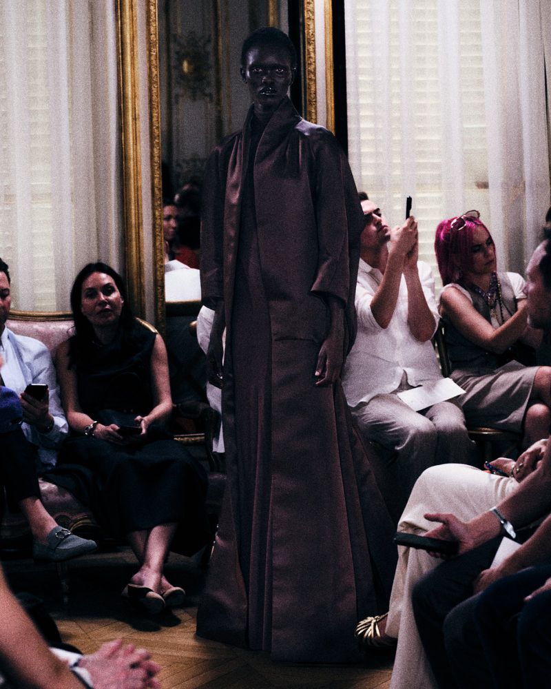 Model wearing a dark brown coat and long dress standing before seated audience members indoors