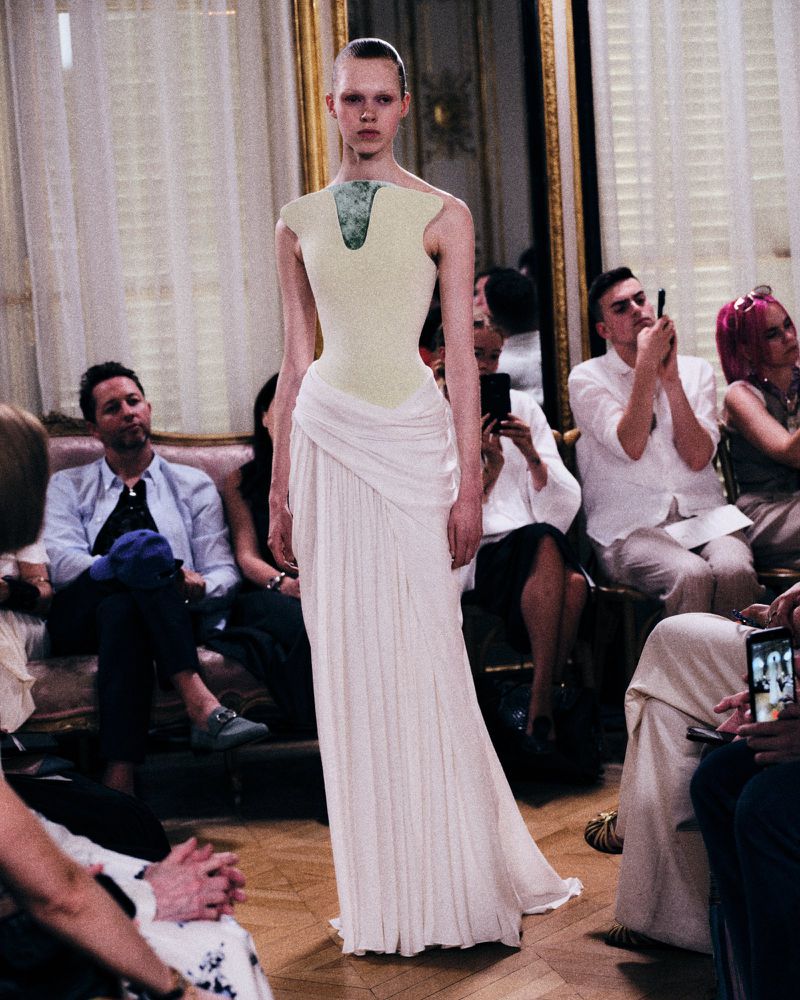 Model walking the runway in a pale green and white sculptural haute couture gown during a fashion presentation.