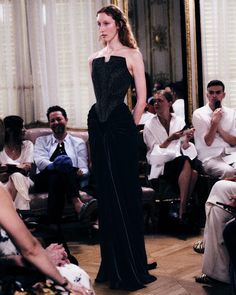 Model in a structured black strapless evening gown walking down a runway before seated guests in an interior salon.