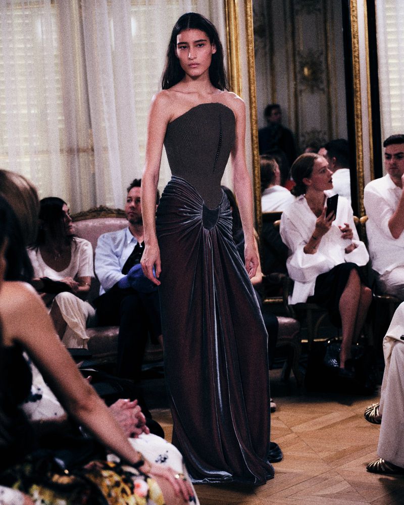 Model walking a runway wearing a strapless fitted dark bodice gown with a draped velvet skirt