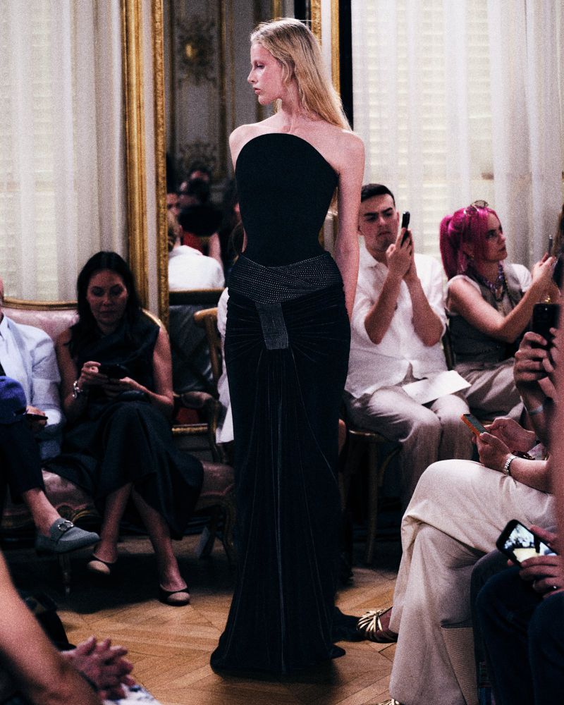 Model in a black strapless gown walking past seated guests holding phones in a salon setting