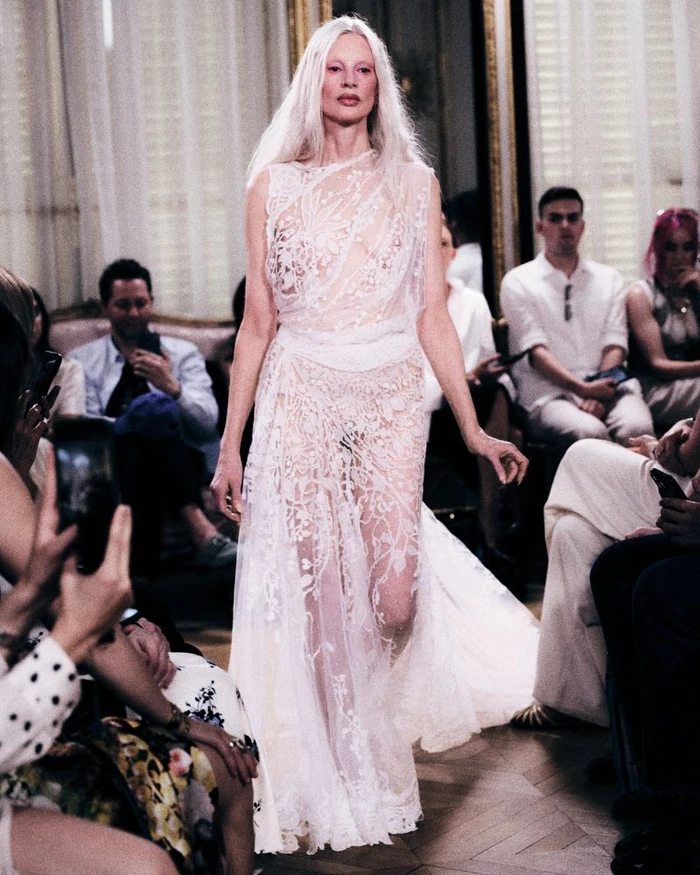 Model with long white hair in a white lace gown walking down a runway during a Standing Ground fashion show.