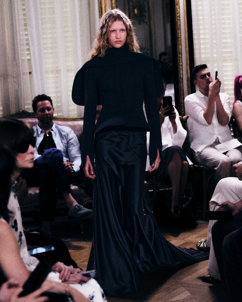 Model walking a runway wearing a dark sculptural turtleneck top and long skirt before seated guests.
