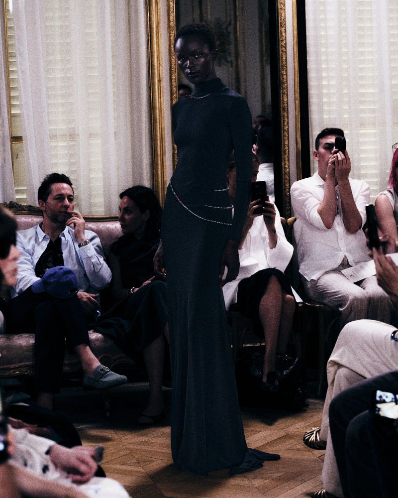 A Black model wearing a long dark gown walking past seated guests in a salon interior for Standing Ground fall couture.