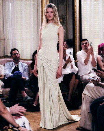 Model in a draped ivory sleeveless gown walking on a runway during a fashion show presentation.
