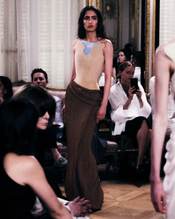 Model wearing a structured sleeveless top and brown patterned maxi skirt walking down a runway before seated guests.