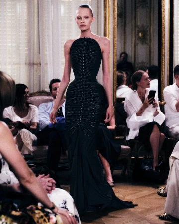 Model in a black floor-length halter gown walking down a runway surrounded by seated audience members