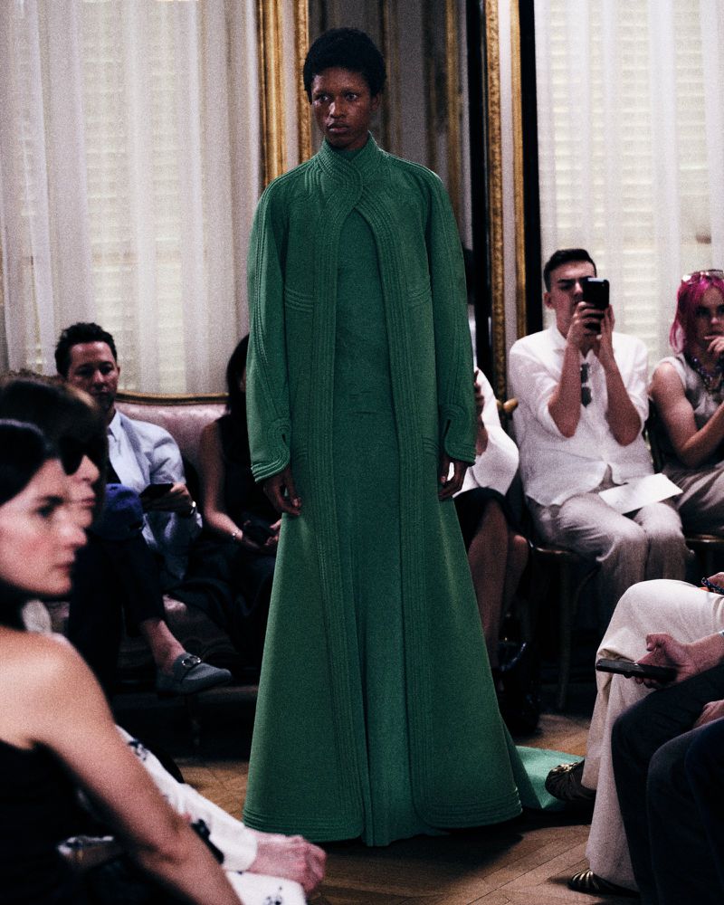 Model wearing a structured emerald green floor-length coat walking past seated audience members during a fashion show.