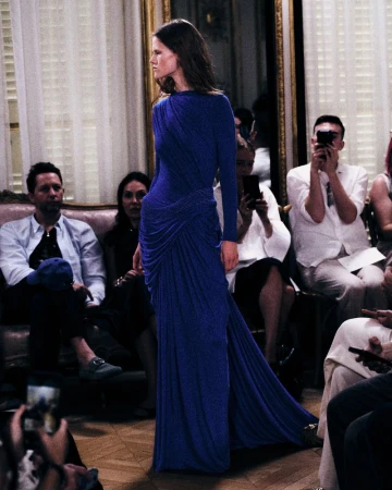 Model in a long blue dress walking on a wooden floor with seated audience members watching.