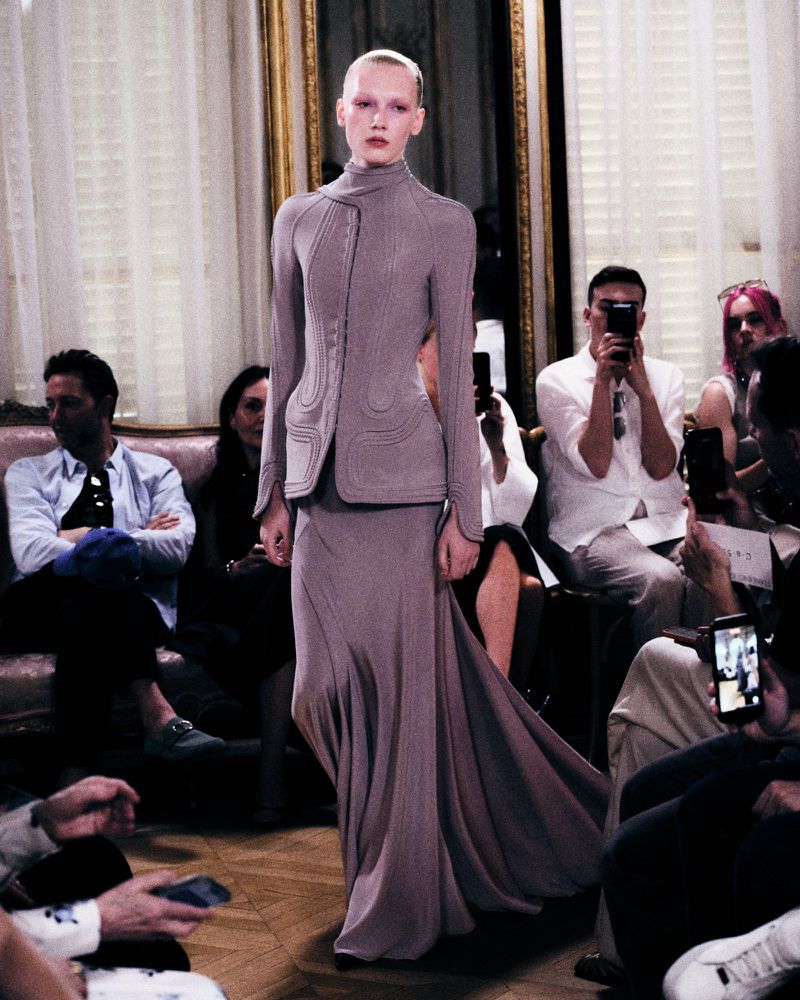 Model walking a runway in a structured grey-mauve jacket and matching long skirt before seated audience members.