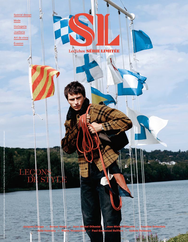 A male model wearing a plaid jacket holding a red rope stands outdoors by a body of water with maritime flags flying behind him.
