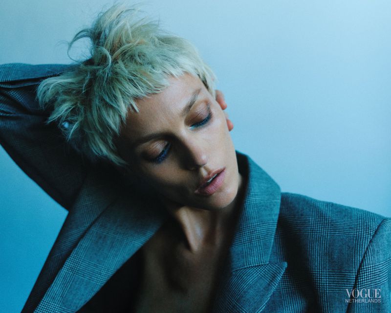 Model Anja Rubik with short blonde hair wearing a grey plaid blazer against a blue backdrop