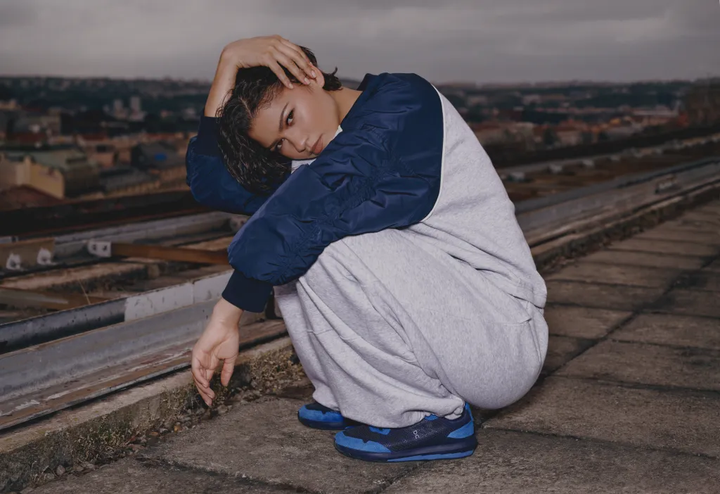 Zendaya crouching on a concrete rooftop wearing a grey and blue tracksuit and blue sneakers against an overcast city skyline
