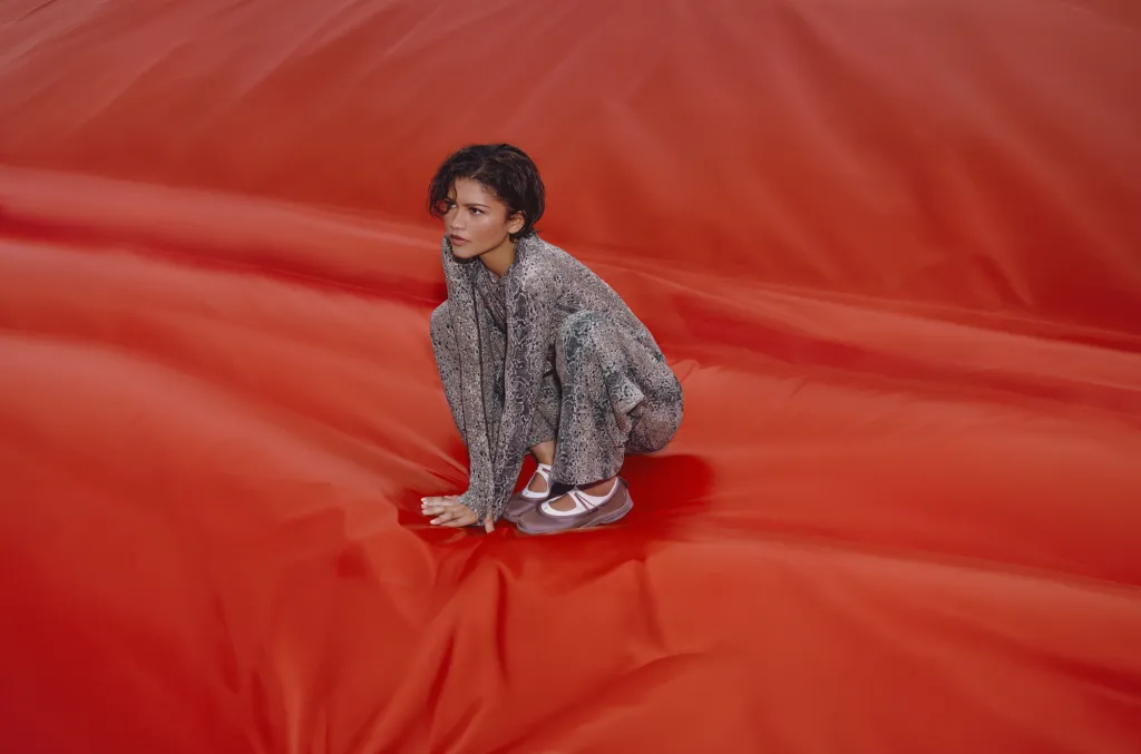 Zendaya crouching on a large textured red fabric backdrop wearing grey patterned apparel and sneakers.
