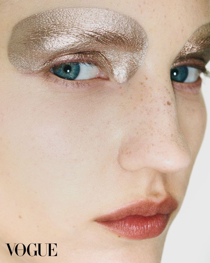 Close-up of a young adult female with dramatic gold eye makeup, light skin, and blue eyes against a stark white background. Vogue logo visible.