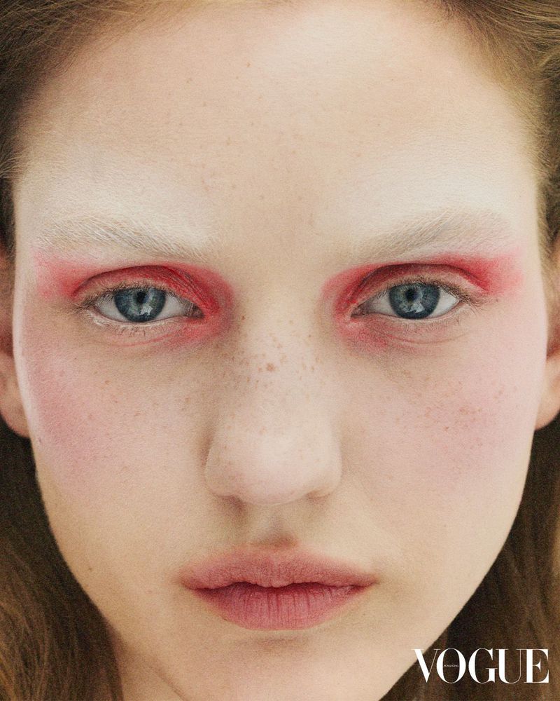 Close-up portrait of a young adult woman with vivid red eye makeup and light hair in a beauty fashion editorial. Vogue logo visible.