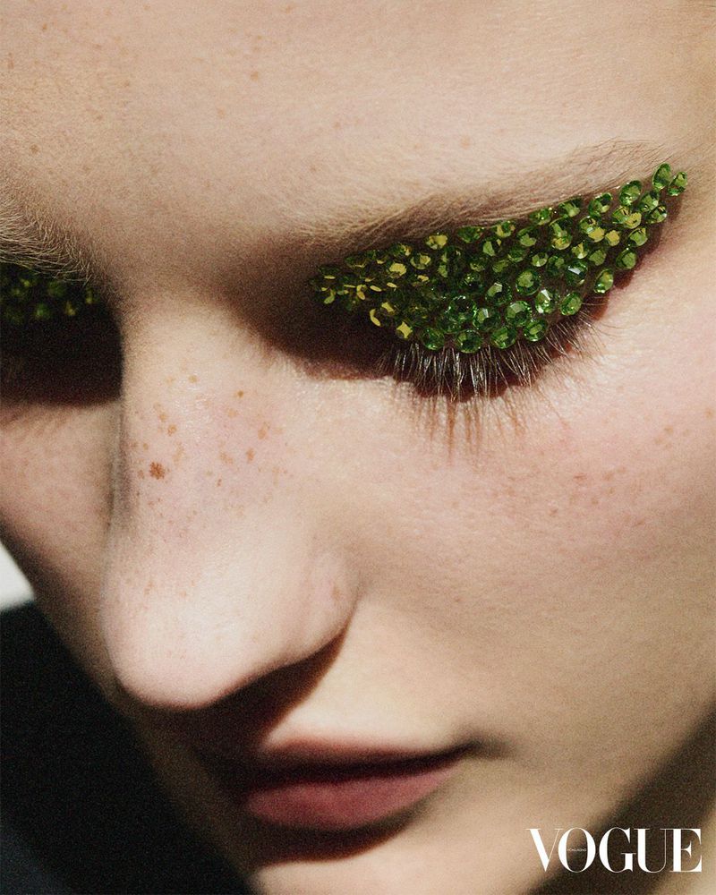 Close-up of a woman's face with green beads applied to her eyebrows and lashes, showcasing a unique beauty and fashion makeup look.